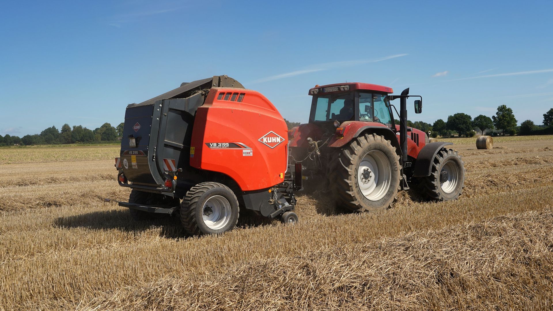 Eine gelbe Rundballenpresse steht neben einem Traktor auf einem grasbewachsenen Feld; eine Person steht in der Nähe unter Bäumen.