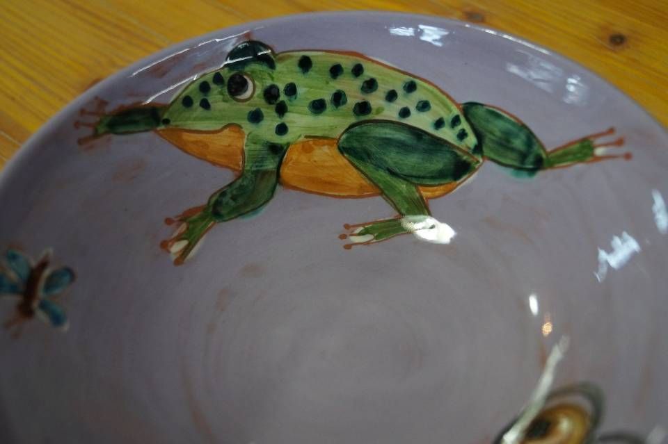 Bemalter grüner Frosch mit schwarzen Punkten auf einem weissen Keramikteller vor einem hölzernen Tischhintergrund
