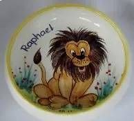 Ein gezeichnetes Löwenjunges mit der Aufschrift „Raphael“ sitzt auf einem weissen Teller mit gelbem Rand im Gras und zwischen Blumen.