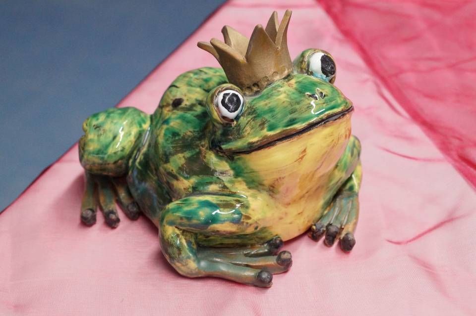 Grüne Keramikfroschfigur mit goldener Krone vor rosa Stoffhintergrund