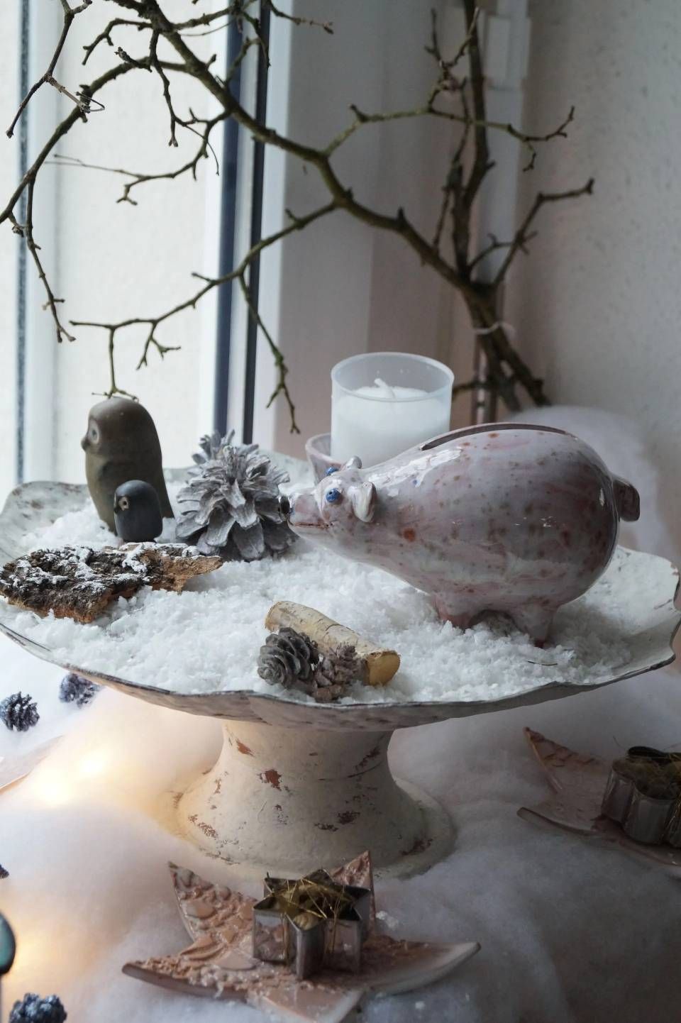 Eine mit Schnee bedeckte Tischplatte mit Tannenzapfen, einer Muschel und einer Kerze auf einem Sockel in der Nähe eines Fensters