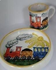 Bemalter Keramikbecher und -teller mit buntem Zugmotiv, Sonne und der Botschaft „Frohe Weihnachten“.