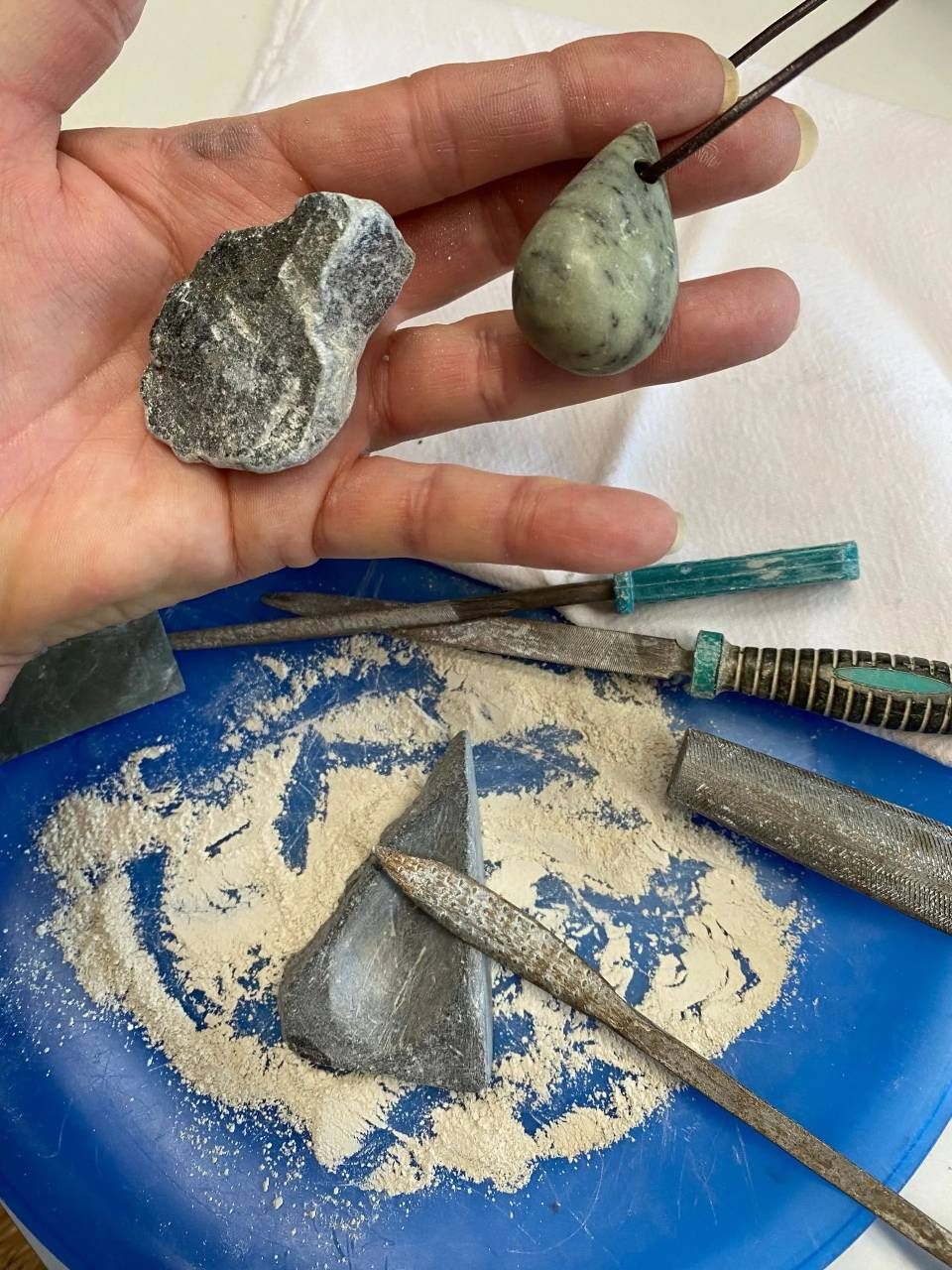 Eine Hand hält zwei Steine ​​über einem blauen Tablett mit Sand und einigen Metallwerkzeugen auf einem Tisch.