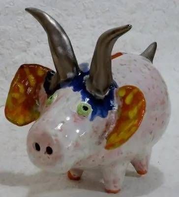 Keramikfigur eines rosa Schweins mit grossen Hörnern, blauen Akzenten und orangefarbenen Ohren auf grauem Untergrund