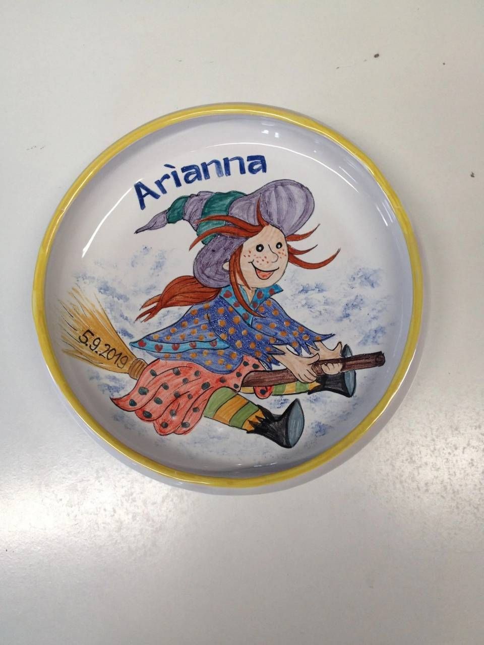 Ein runder Anstecker mit der Aufschrift „Arianna“ und einer lächelnden Comic-Hexe, die auf einem Besen reitet, mit gelbem Rand.