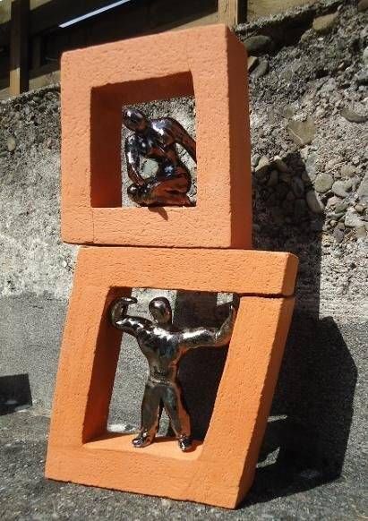 Zwei orangefarben umrahmte, quadratische Skulpturen, schief übereinander gestapelt, jede mit einer kleinen Metallfigur, vor einem felsigen Hintergrund.