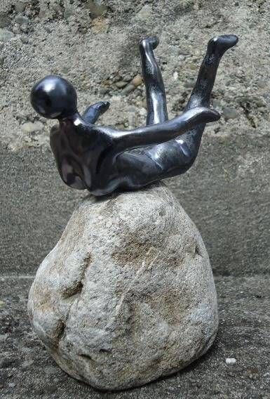 Kleine dunkle Skulptur einer liegenden Figur, die im Freien auf einem grossen Felsen balanciert.