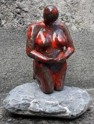 Eine rot glänzende, kniende Skulptur auf einem rauen, grauen Steinsockel vor einer strukturierten Wand.