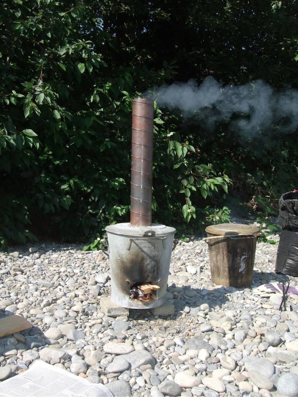 Kleiner Holzofen mit rauchendem Schornstein auf felsigem Untergrund im Freien nahe Bäumen