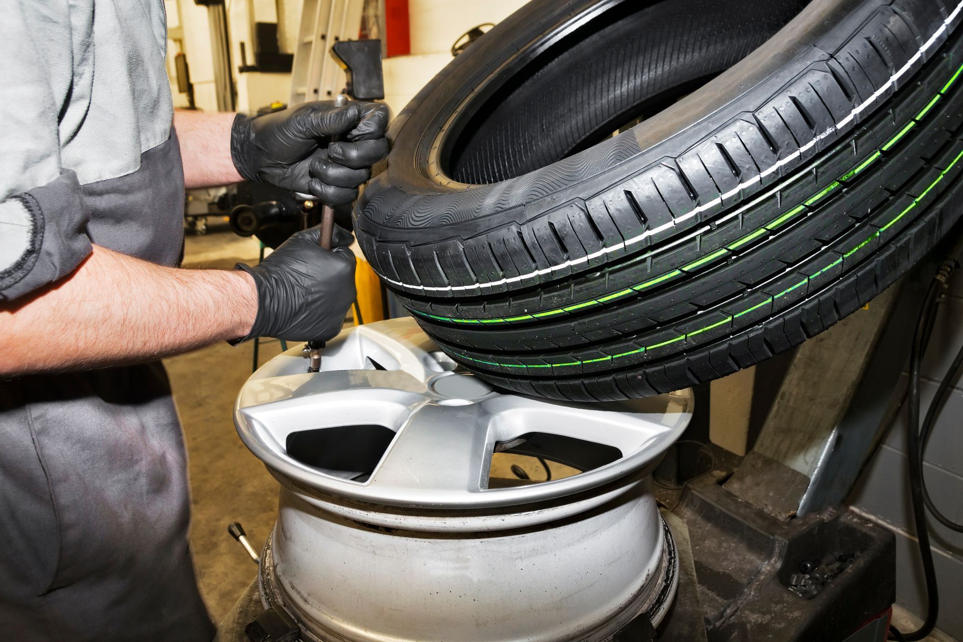 Mécanicien montant un pneu sur une jante argentée à l'aide d'une machine à pneus ; gants noirs, environnement intérieur.