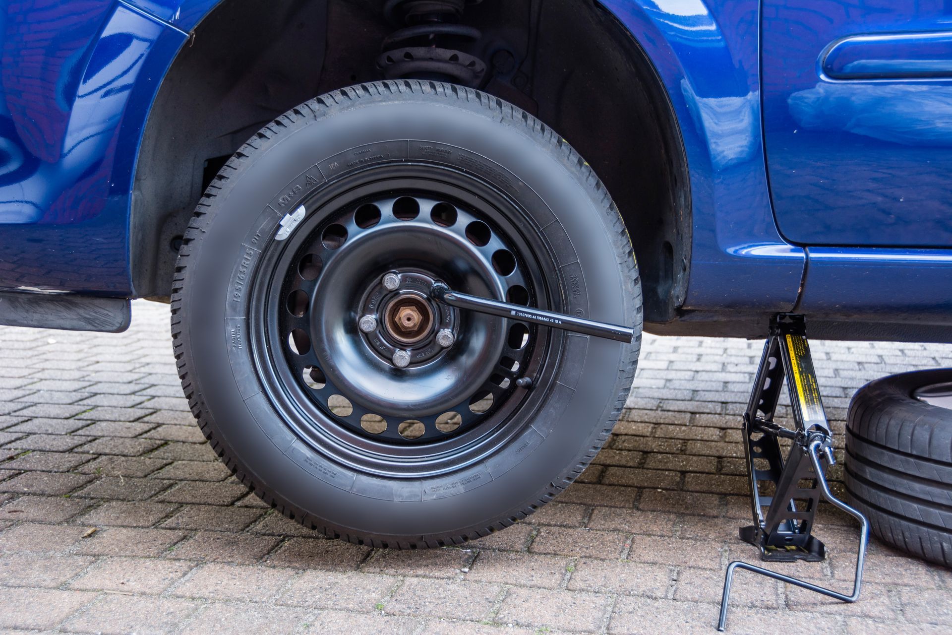 On change un pneu bleu de voiture avec un cric, la roue de secours est au sol et on utilise une clé de roue.