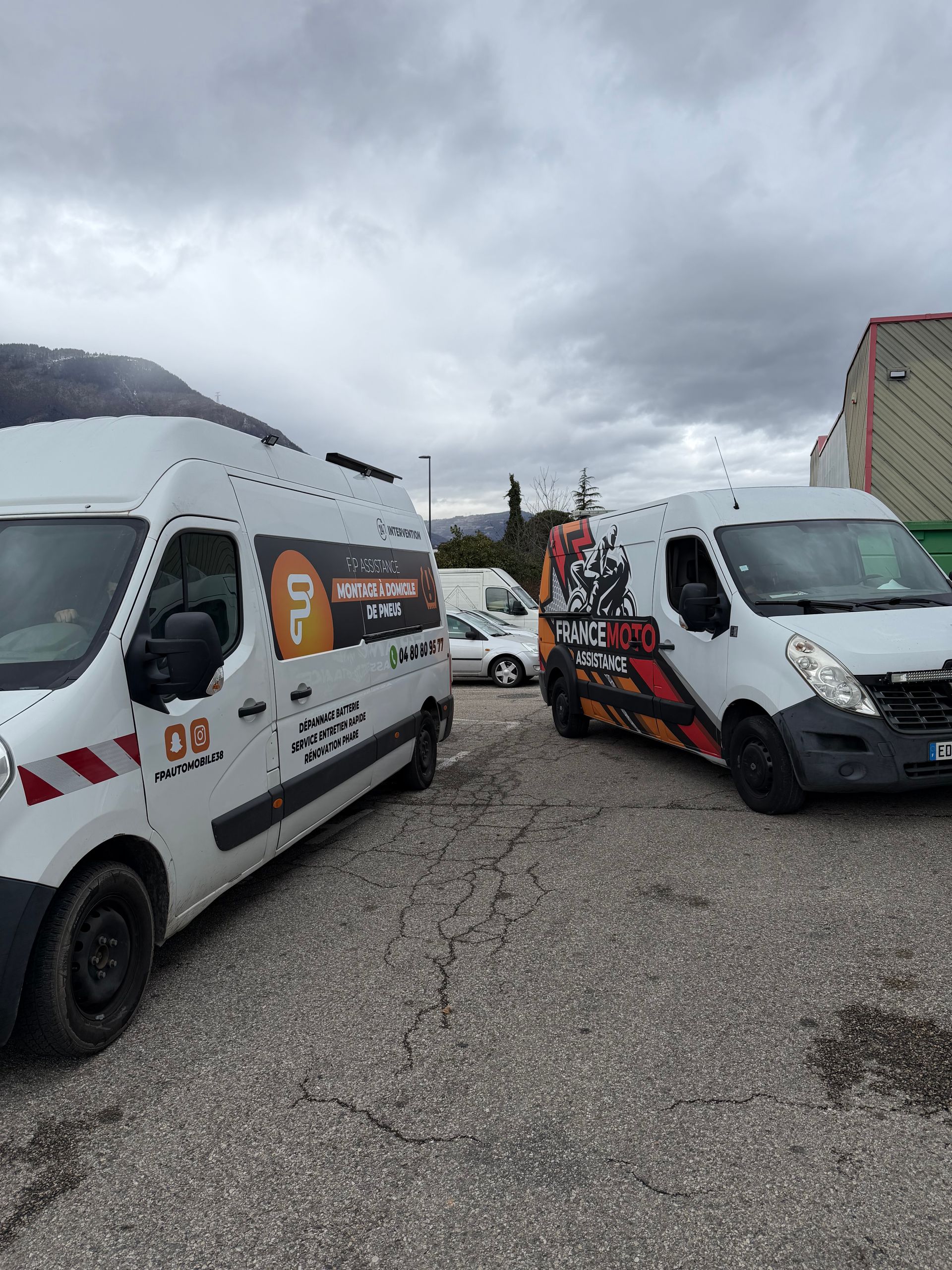 Deux fourgonnettes commerciales blanches arborant des logos sont garées sur un terrain gravillonné.