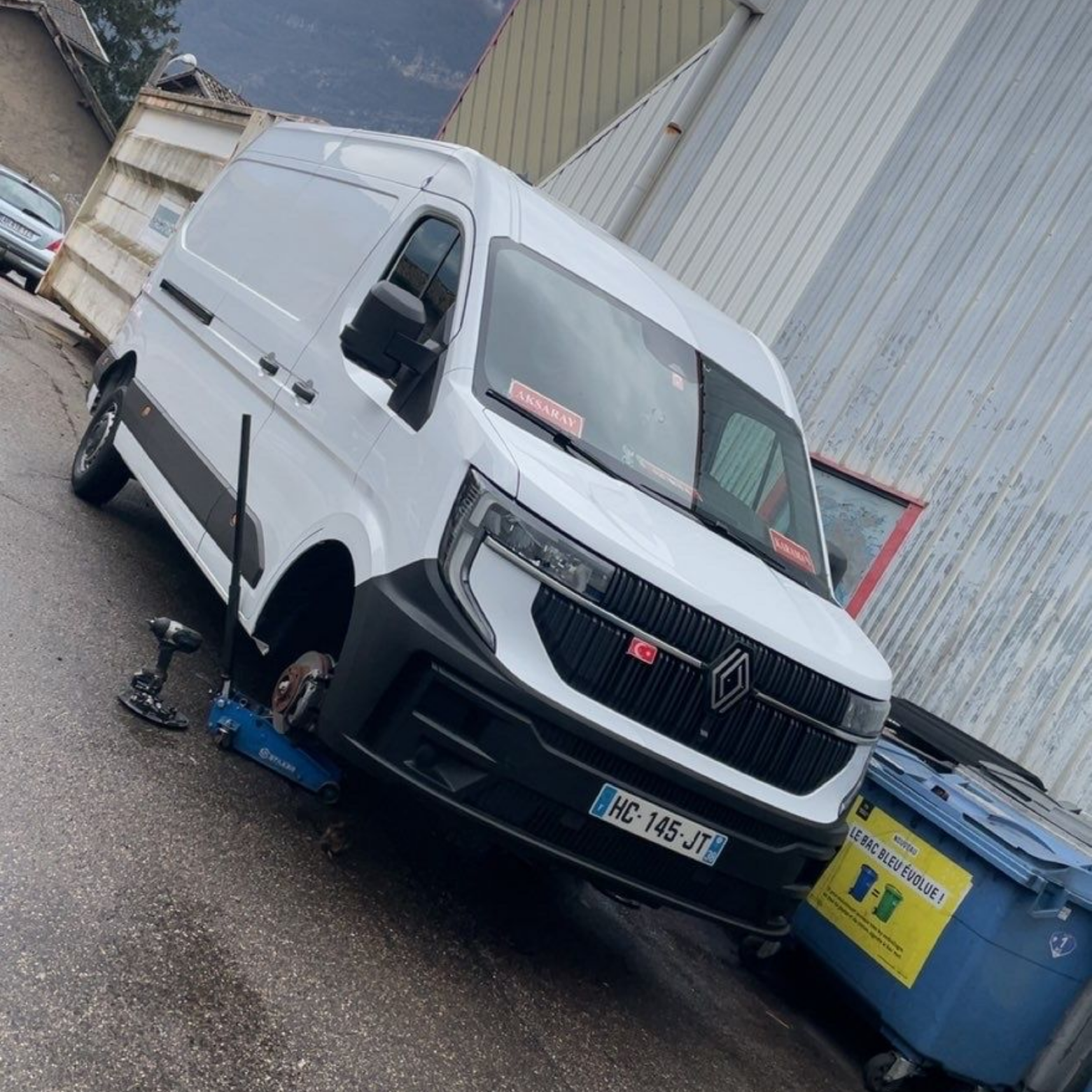Une camionnette de livraison Renault blanche est calée par un cric hydraulique bleu pour des travaux d'entretien.
