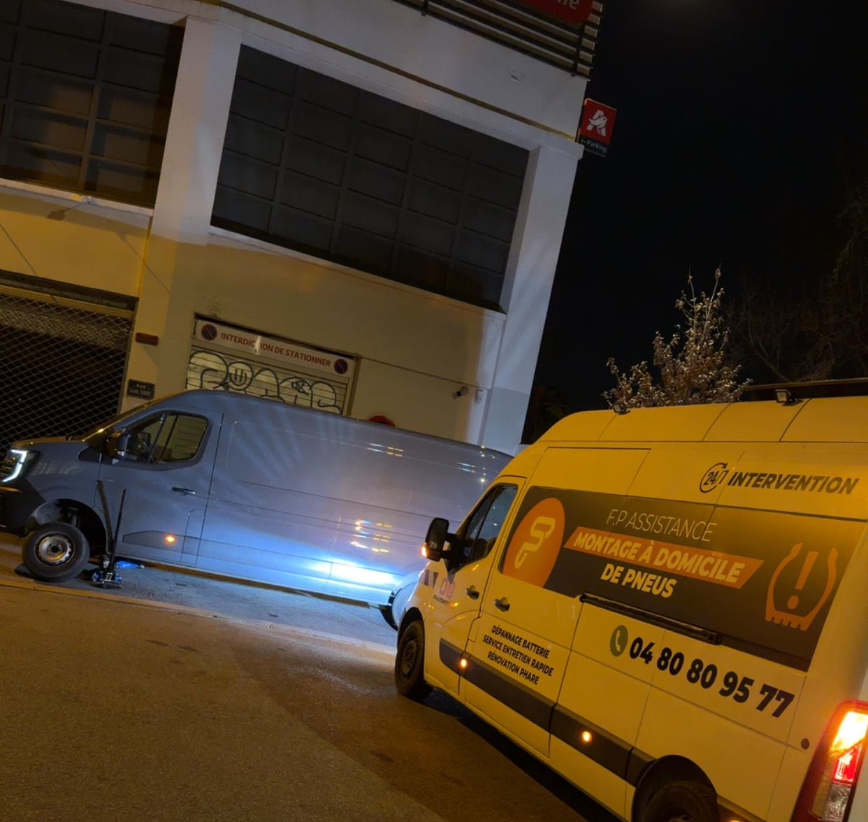 Une camionnette de dépannage pneumatique stationnée de nuit à côté d'une camionnette grise dont la roue avant a été démontée pour assistance routière.