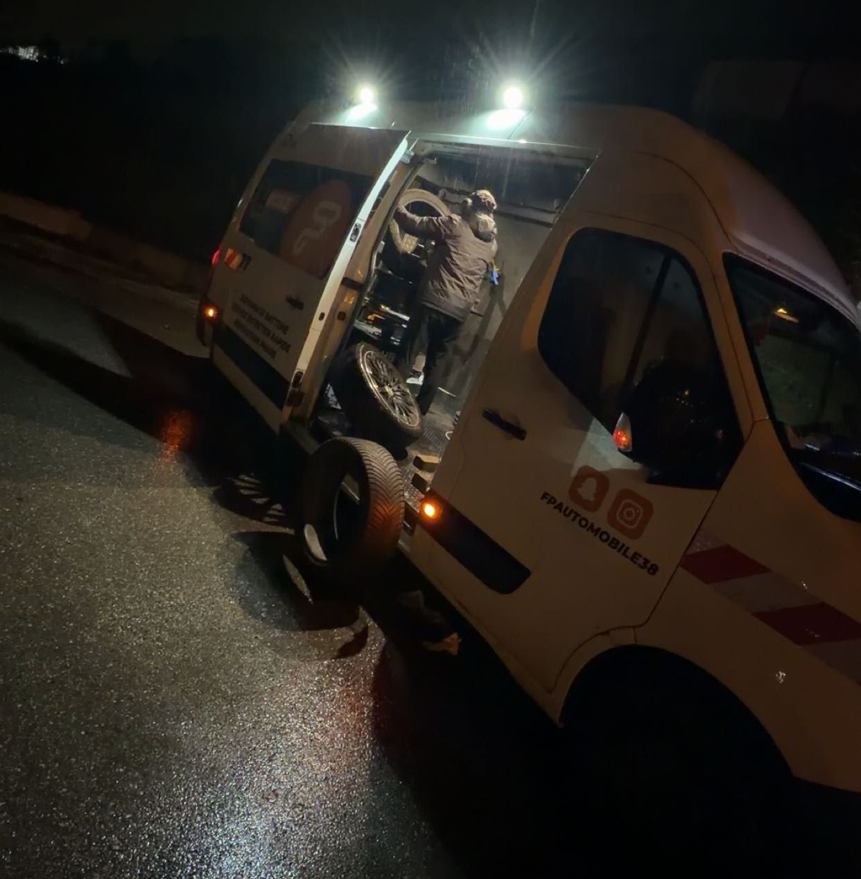 La nuit, un dépanneur charge des pneus à l'arrière d'une camionnette blanche sur une route sombre et mouillée.
