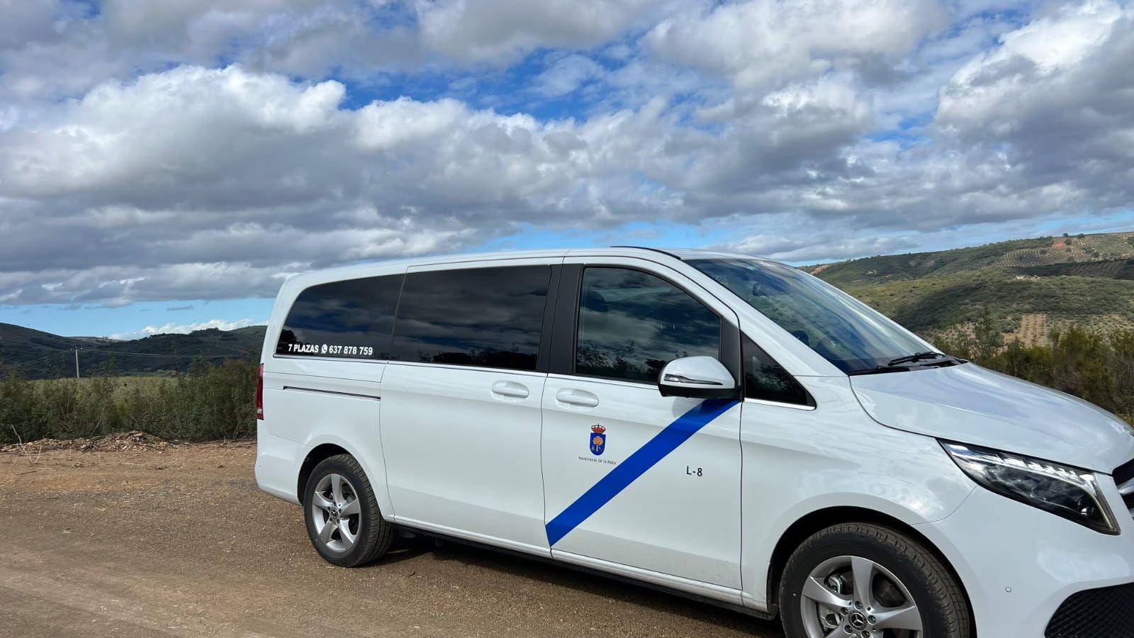 Furgoneta taxi blanca en un camino de tierra, franja azul, cielo nublado y colinas al fondo.