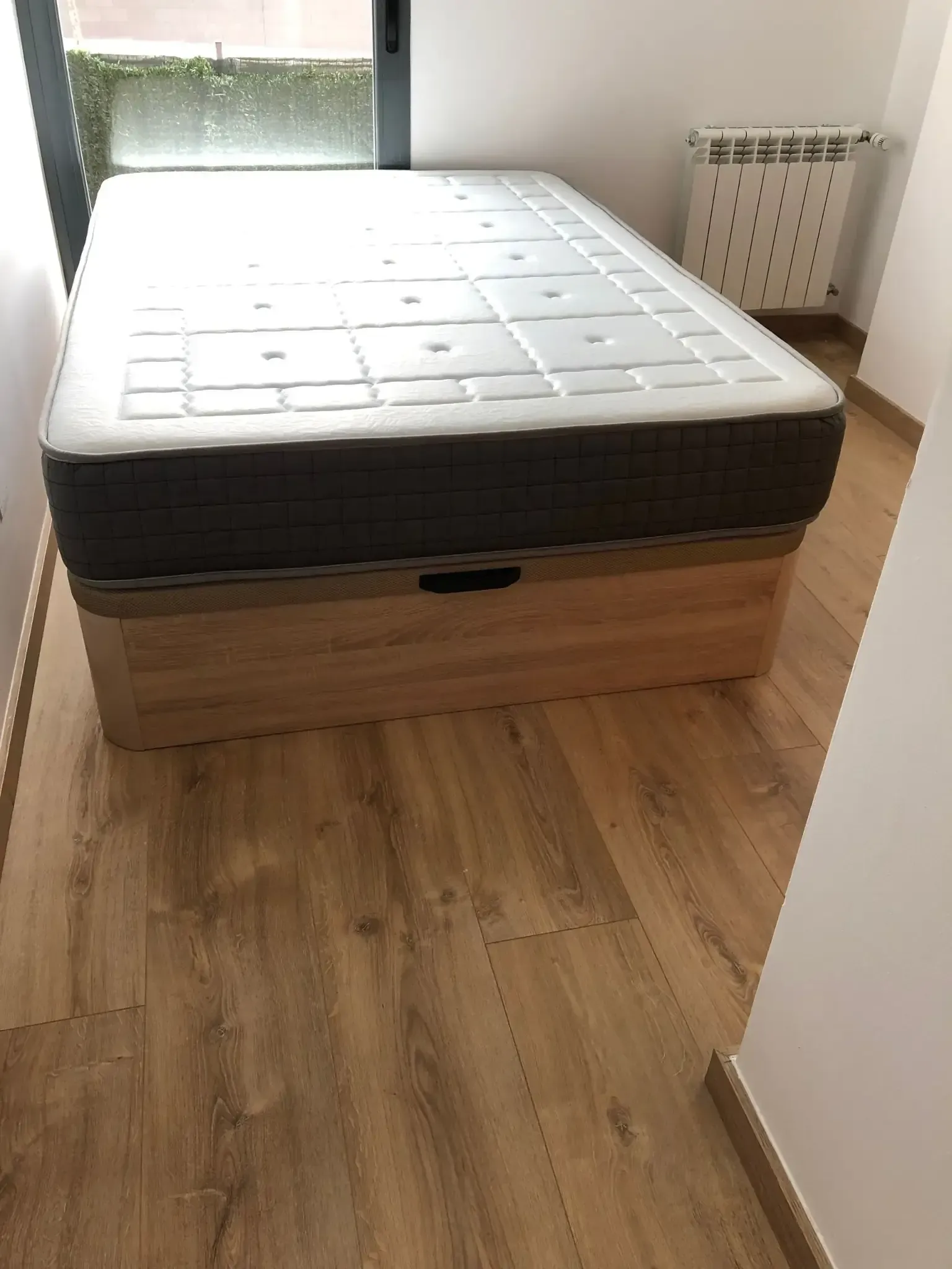 Cama con colchón gris sobre una base de almacenamiento de madera clara, en una habitación con suelo de madera.