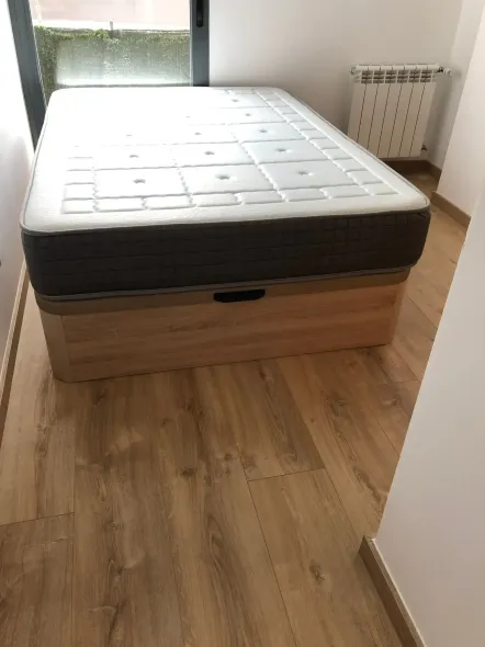 Cama con colchón gris sobre una base de almacenamiento de madera clara, en una habitación con suelo de madera.