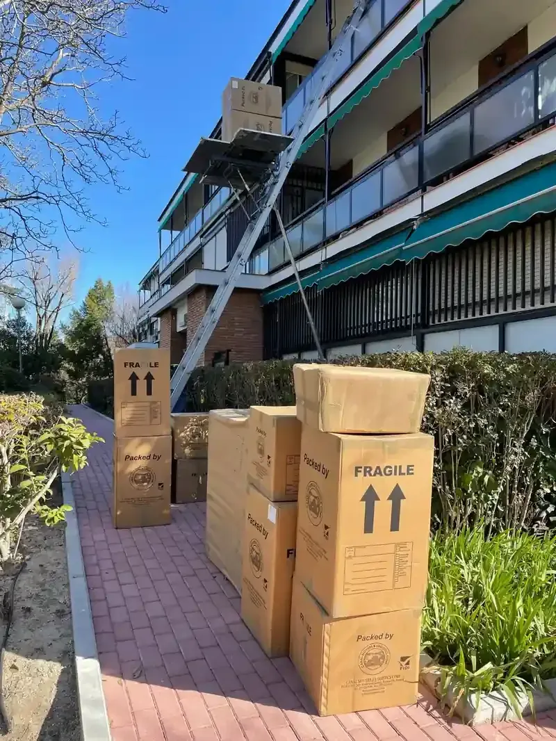 Cajas de mudanza apiladas en la acera, con un ascensor que da acceso al edificio de apartamentos. Día soleado.