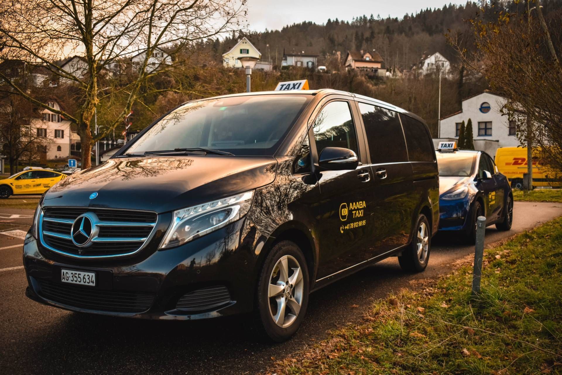 Ein schwarzes Mercedes-Taxi parkt am Strassenrand; im Hintergrund sind Häuser und Hügel zu sehen.