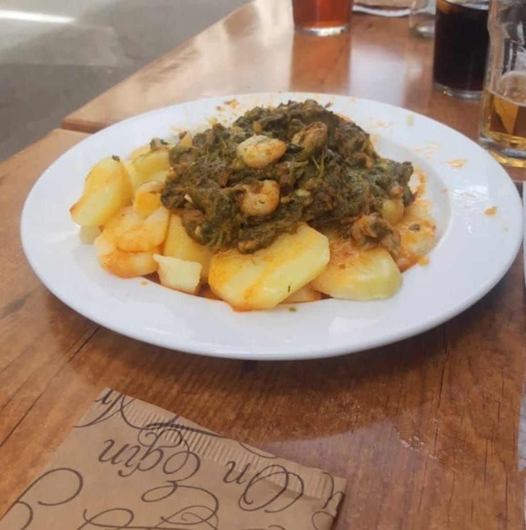 Plato de patatas con salsa verde sobre una mesa de madera, junto a una servilleta y unos vasos.