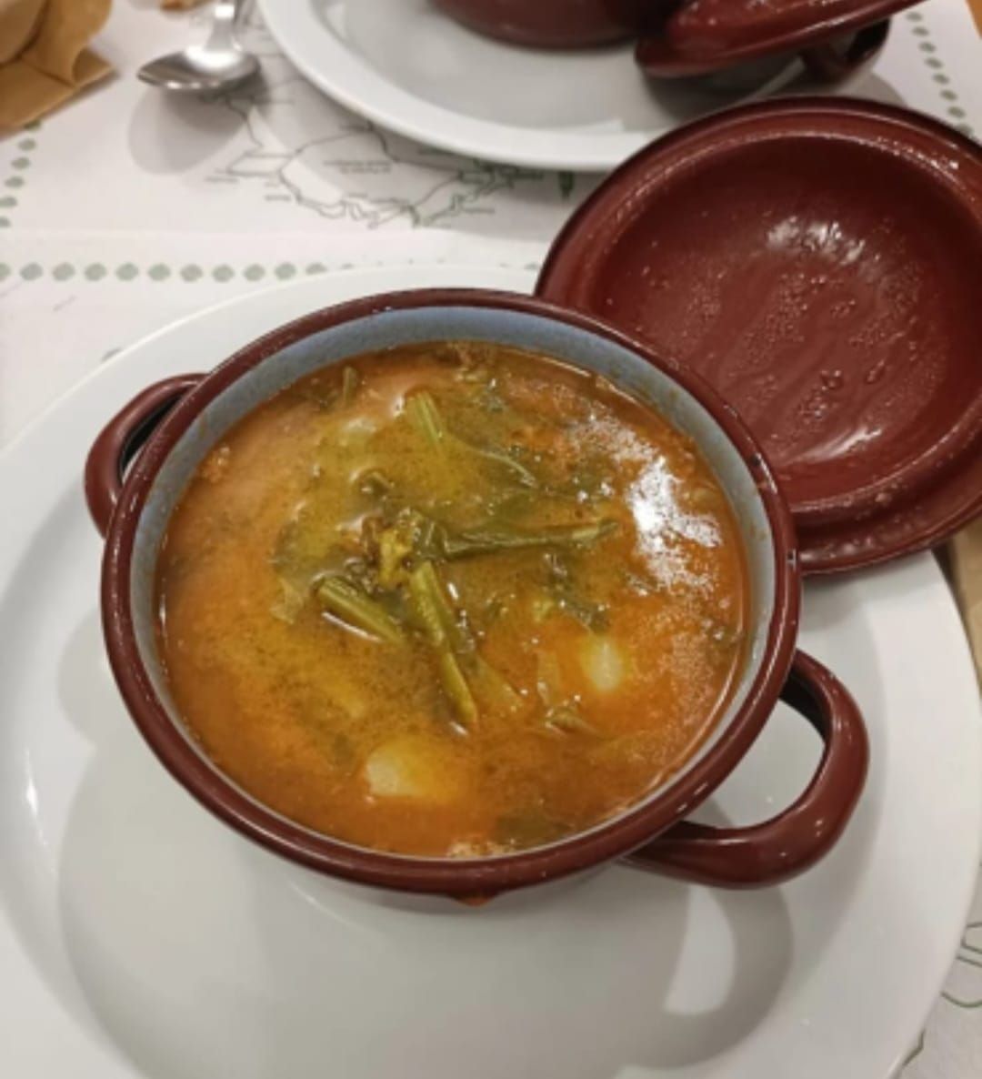 Sopa en un tazón marrón con asa y tapa; verduras, plato blanco, mantel.