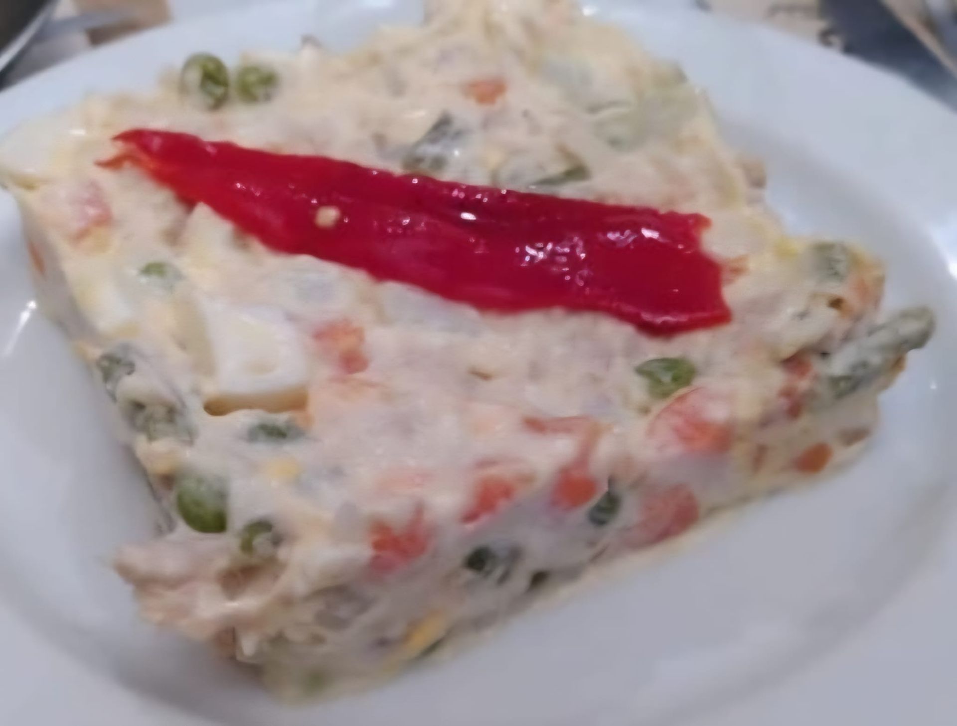 Porción cuadrada de ensalada de patatas cubierta con una tira de pimiento rojo en un plato blanco.