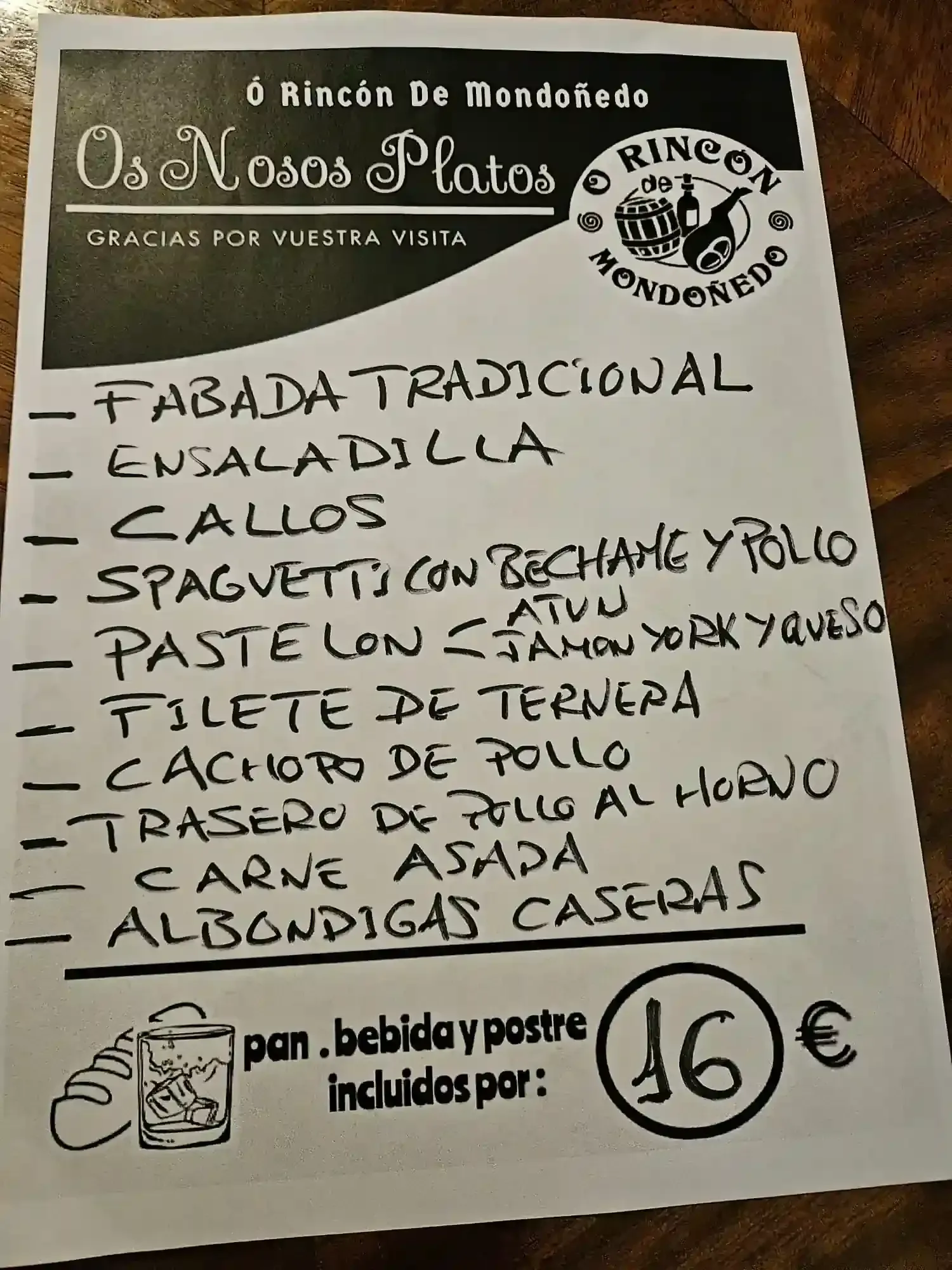 Menú escrito a mano con tinta negra sobre papel blanco, con una lista de platos por 15 € que incluye pan, bebida y postre.