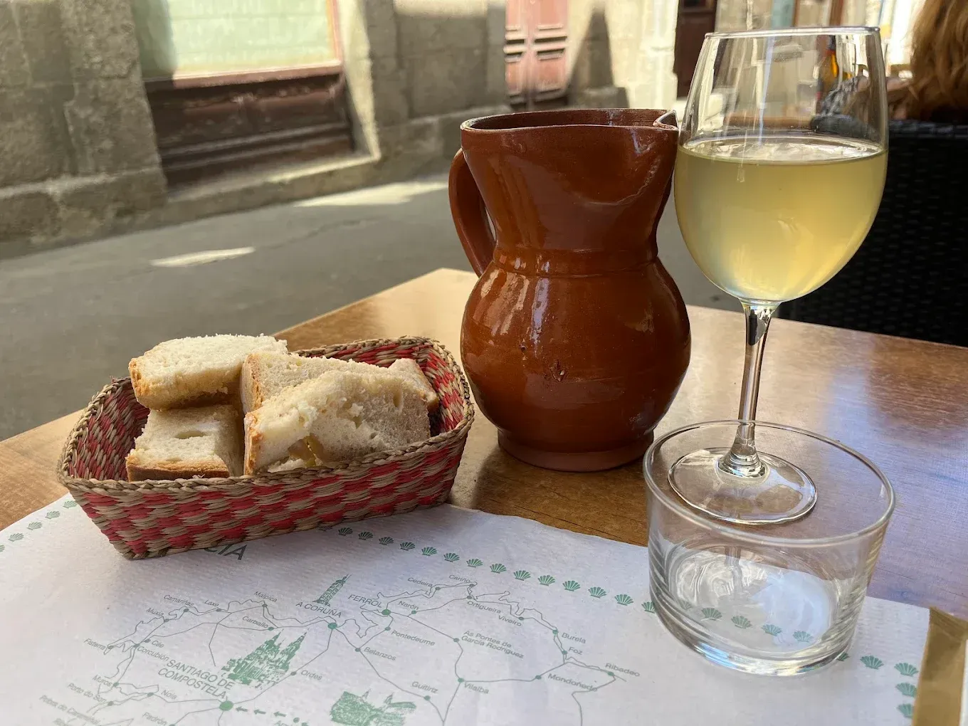 Cesta de pan, jarra de barro, copa de vino blanco y vaso de agua sobre una mesa. Ambiente exterior.