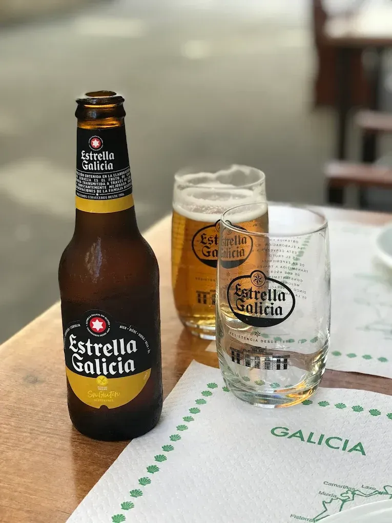 Una botella y dos vasos de cerveza Estrella Galicia, parcialmente llenos, están sobre una mesa.