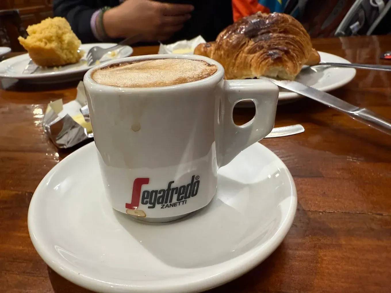 Taza de café Segafredo en un platillo, croissant y muffin sobre una mesa de madera.