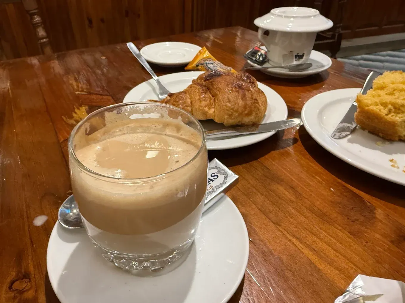 Café, cruasán y pastel sobre una mesa de madera, en un ambiente de cafetería.
