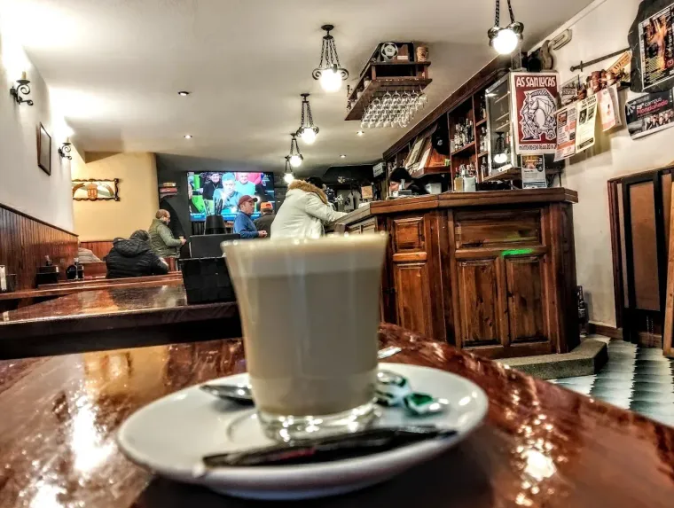 Café servido en un platillo en un bar-cafetería. El interior es de madera y se ven un camarero y clientes.