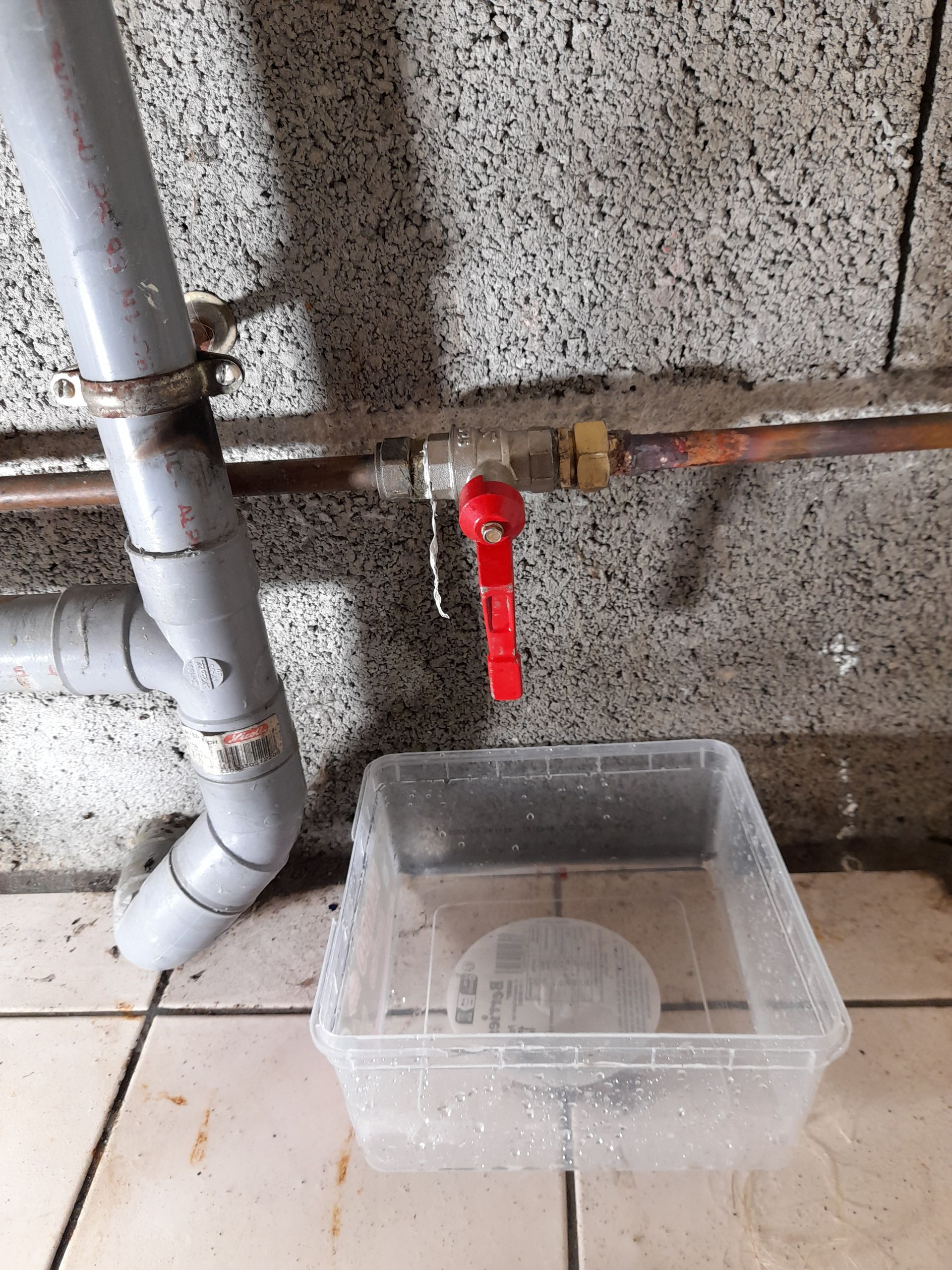 Un plombier serre un tuyau métallique sous un évier avec une clé rouge ; de l’eau goutte du raccord.