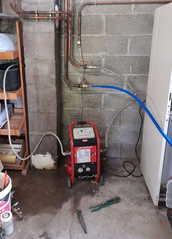 Au sous-sol, une buanderie avec un poste à souder rouge, un mur en parpaings, un tuyau bleu à côté de canalisations et un appareil électroménager blanc.