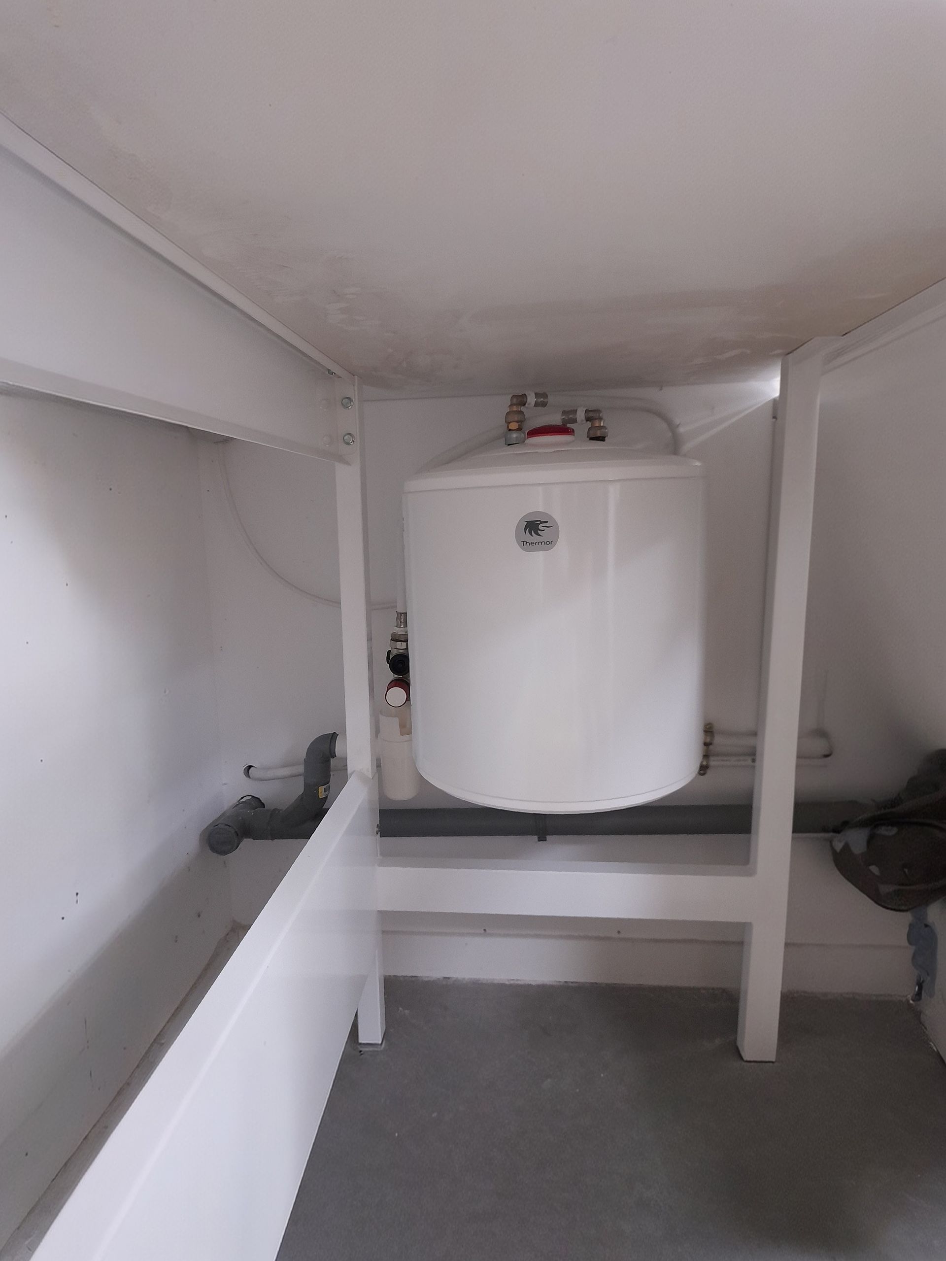 Chauffe-eau blanc installé dans un meuble avec des tuyaux et un sol gris.