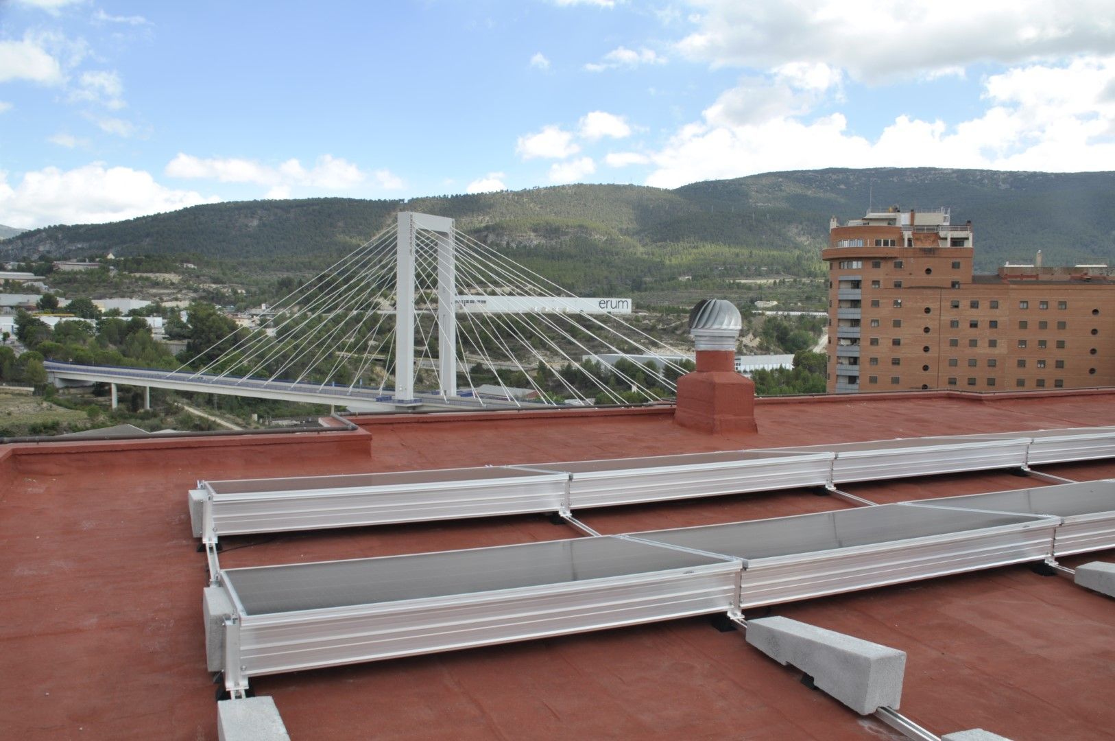 Vista de un puente atirantado y un edificio con techo rojo contra un fondo de montañas bajo un cielo nublado.
