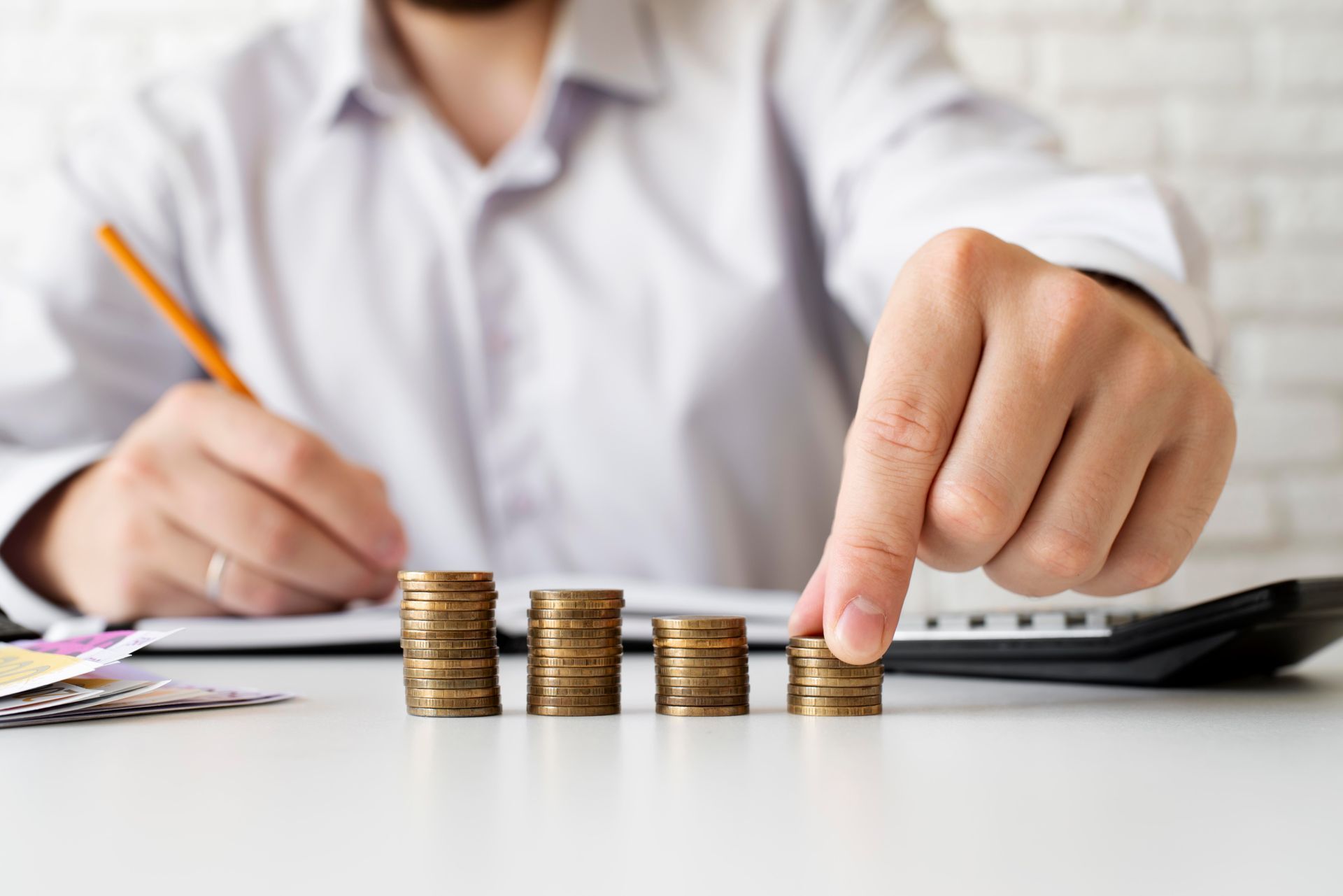 Persona apilando monedas, señalando un pequeño montón. Calculadora, documentos y lápiz sobre la mesa.