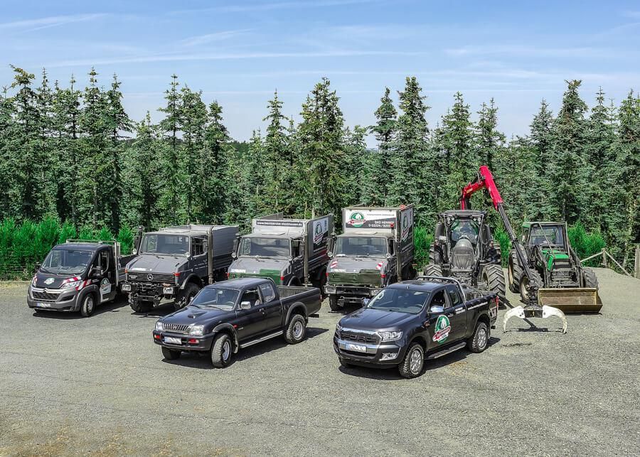 Eine Gruppe von Lastwagen und Lieferwagen parkt auf einem Schotterplatz vor einem Waldstück.
