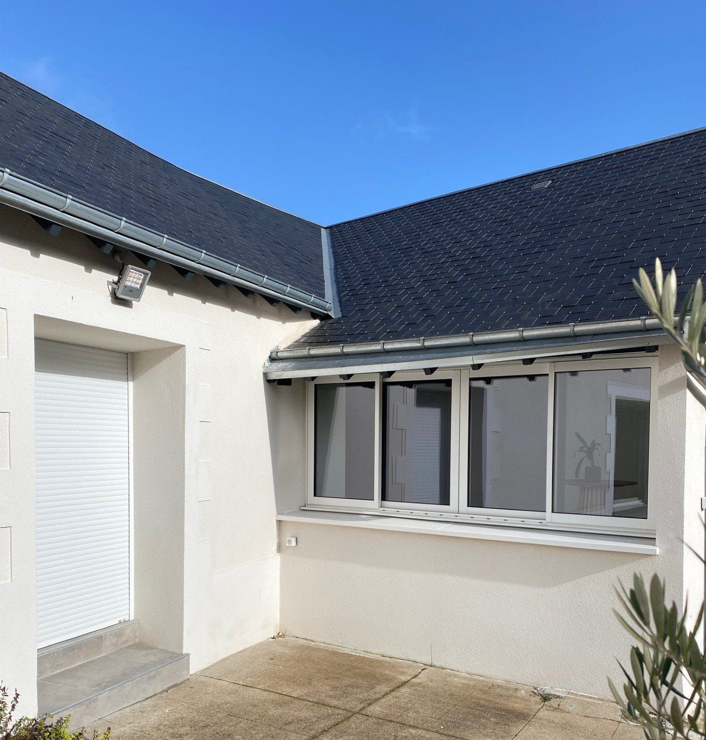 Cour intérieure d'une maison blanche au toit bleu foncé, porte à volets et grandes fenêtres sous un ciel d'un bleu limpide