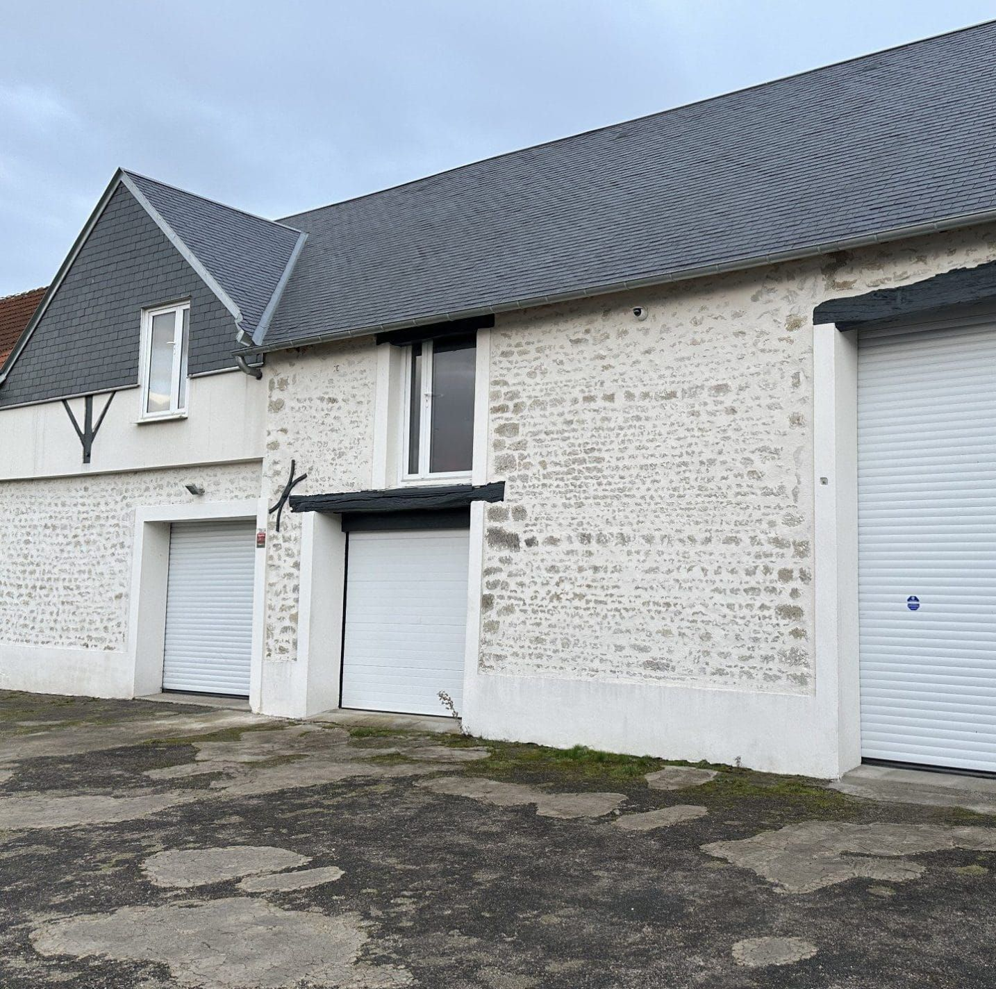 Bâtiment en pierres blanches avec un toit de tuiles grises et trois portes de garage blanches sous un ciel couvert