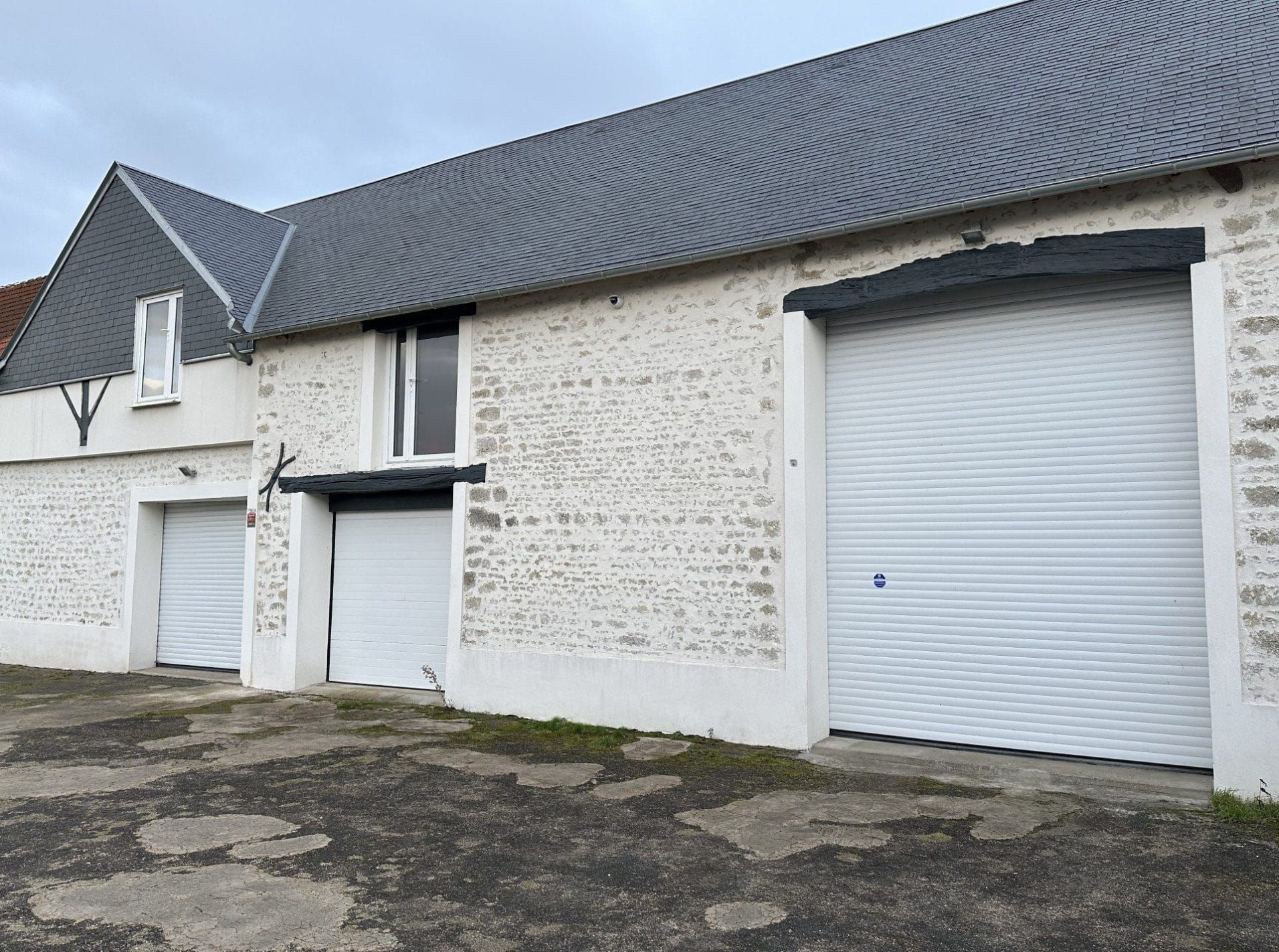 Bâtiment en pierres blanches avec toit gris, portes de garage et petite entrée sur un terrain asphalté fissuré