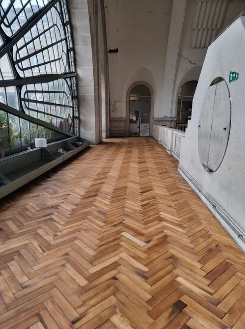 Long couloir au sol en bois à chevrons avec fenêtres cintrées et murs dans un bâtiment historique.