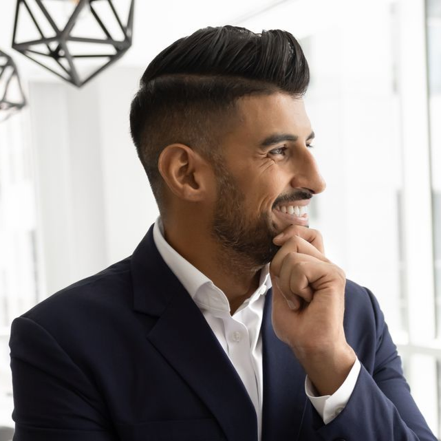 Homme en costume bleu marine, souriant, regardant sur le côté, la main sur le menton. Intérieur lumineux.