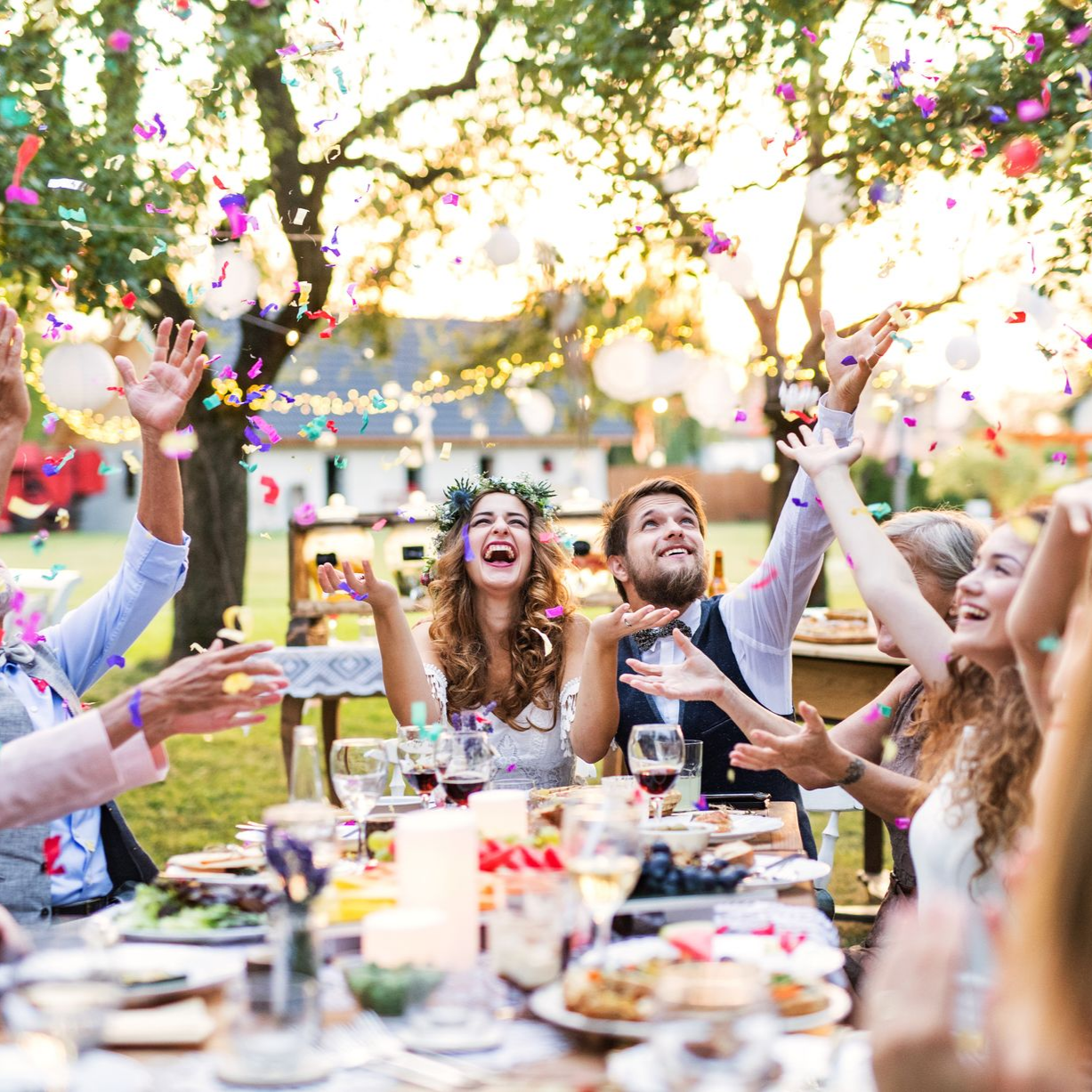 Des invités lancent des confettis et applaudissent lors d'une fête en plein air. Les mariés sourient à une table.