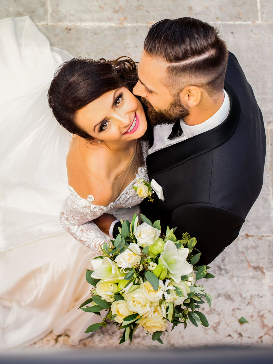 Les mariés s'enlacent, il l'embrasse sur la joue. Elle tient un bouquet.