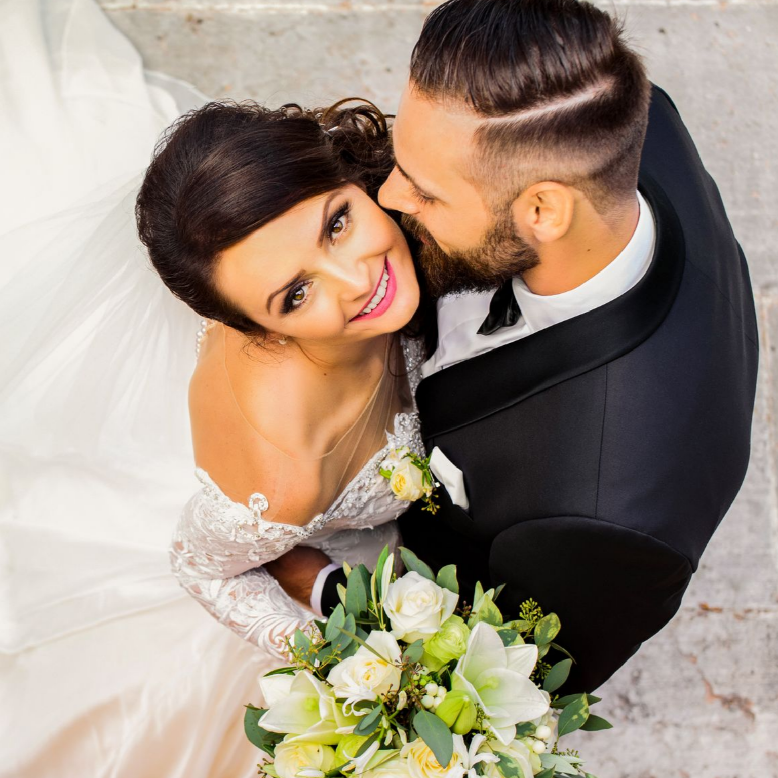 Les mariés s'enlacent le jour de leur mariage, tenant leur bouquet, le marié embrasse la joue de la mariée, vue de dessus.