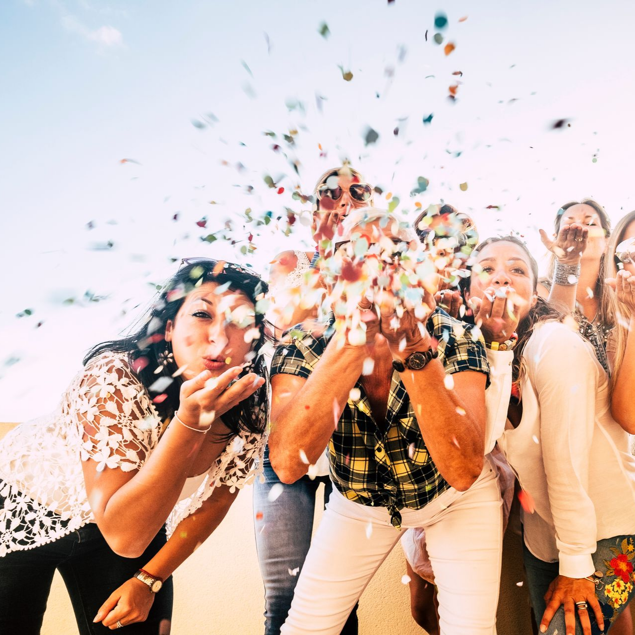 Des personnes soufflent des confettis colorés vers la caméra. En extérieur, sous un soleil radieux. Expressions joyeuses.