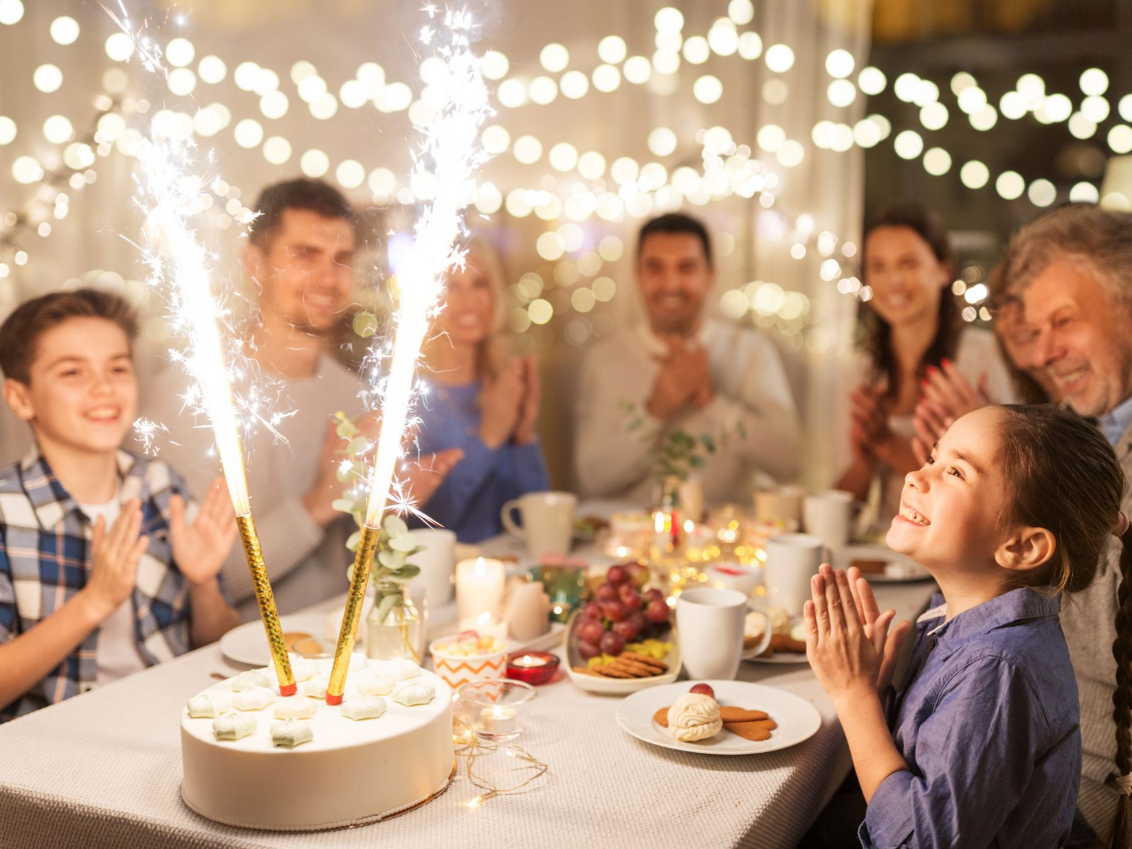 Une famille fête un anniversaire ; un enfant regarde un gâteau orné de cierges magiques ; une table est dressée avec de la nourriture et des lumières sont visibles en arrière-plan.