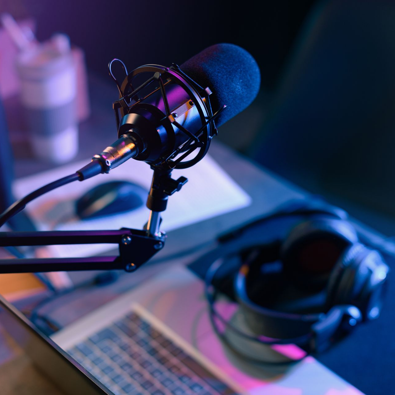 Un microphone est posé sur un bureau, à côté d'un ordinateur portable, d'une souris et d'un casque audio. L'éclairage est violet et bleu.